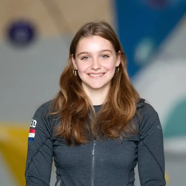 Een jonge vrouw met lang bruin haar, gekleed in een donker jasje met een klein embleem van de Nederlandse vlag, glimlacht naar de camera tegen een wazige, kleurrijke achtergrond.
